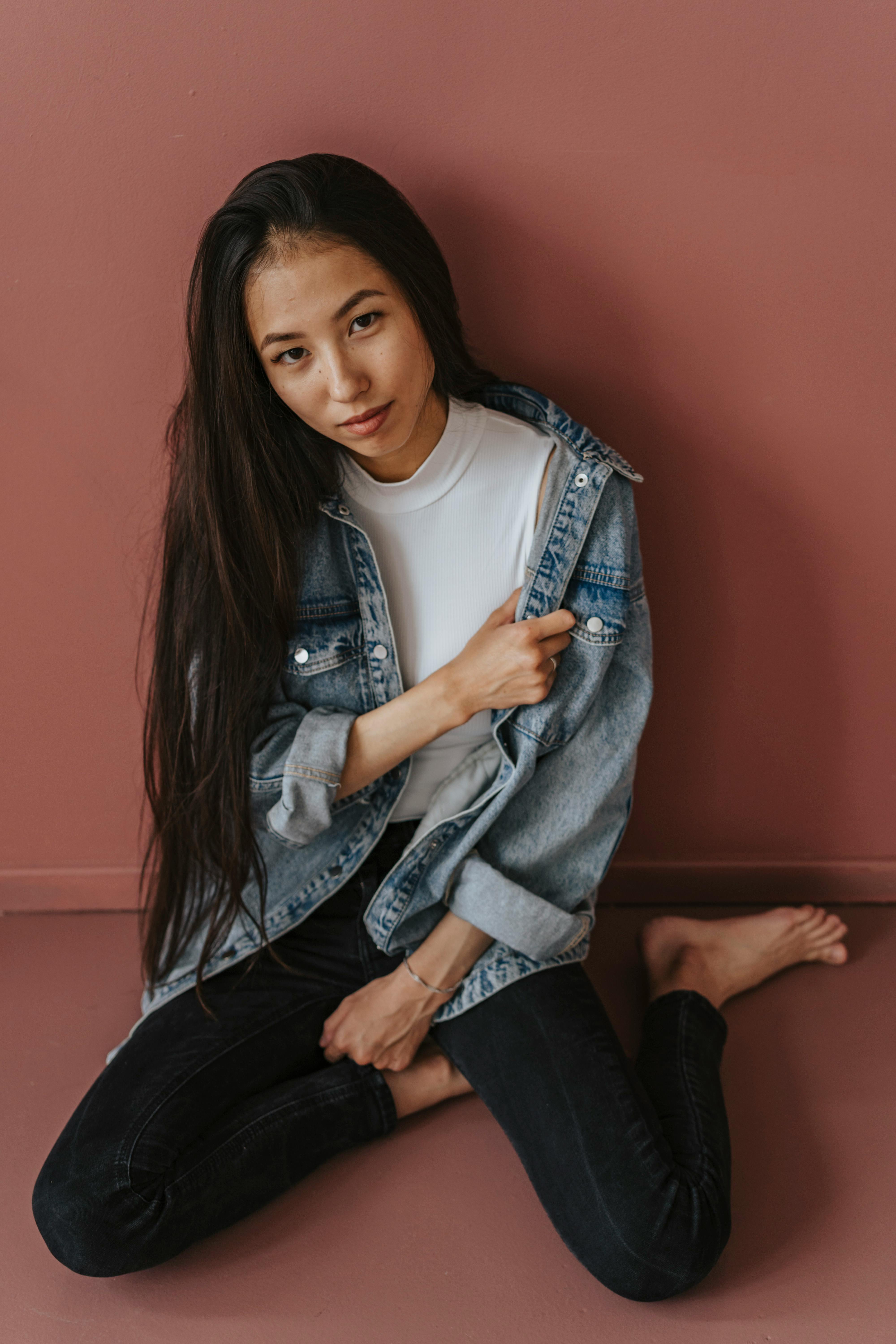 Asian woman posing barefoot in denim jacket against a brown wall.