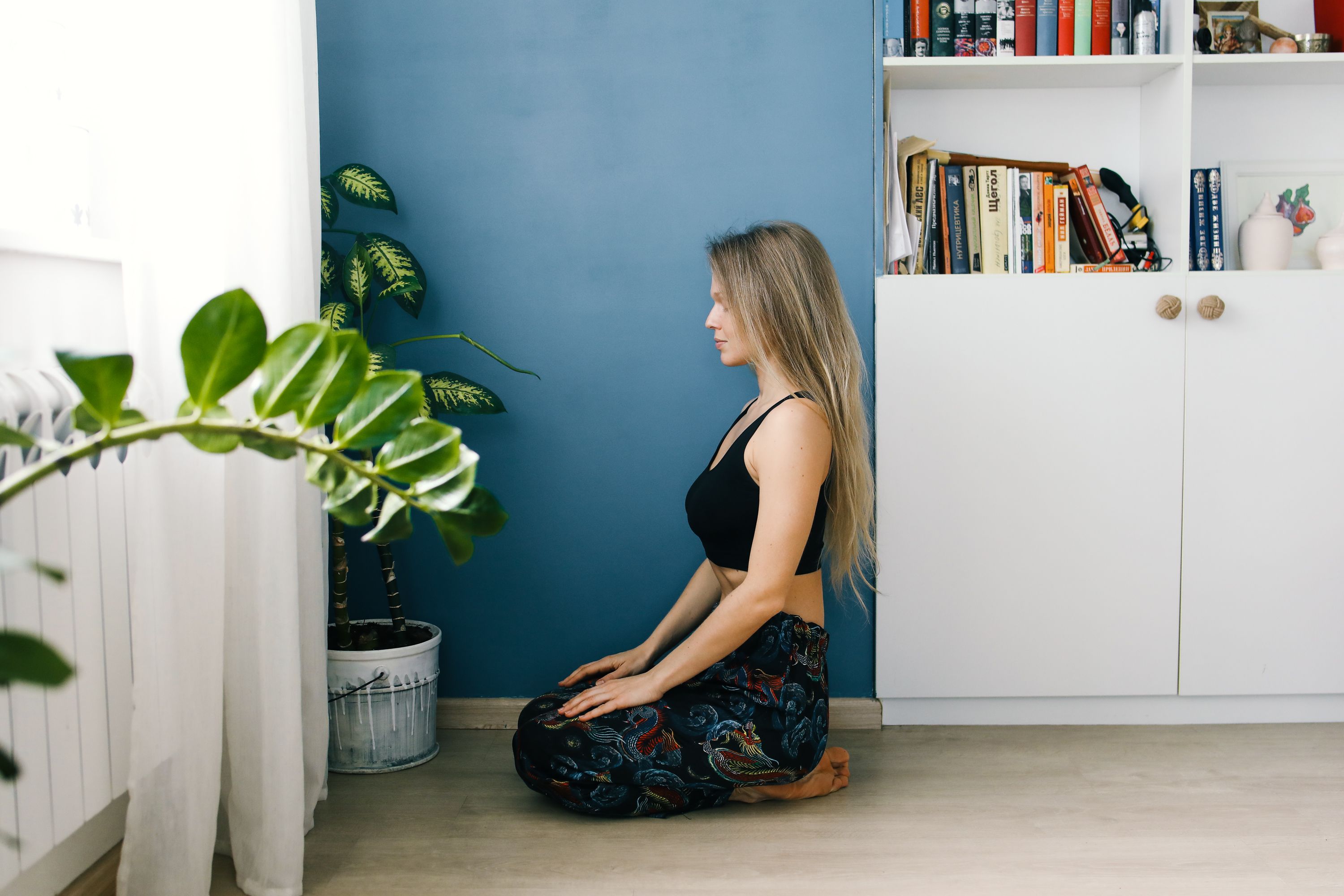Serene woman practicing meditation indoors next to houseplants and a bookshelf.