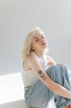 一位金发女子穿着牛仔裤坐着，眨着眼睛，展示了她手臂上的纹身