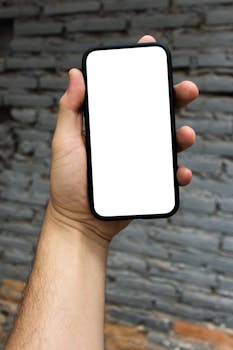 Close up of a hand holding a smartphone with a blank screen, set against a textured brick wall backdrop.