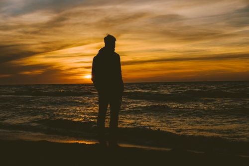 Free A silhouette of a person standing on the beach during a vibrant sunset, creating a serene seascape. Stock Photo