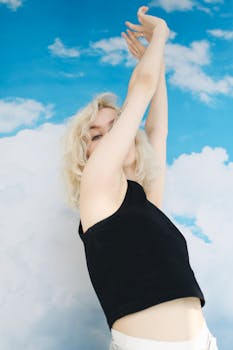 一位年轻的金发女子身穿黑色上衣，在充满活力的蓝天下举起双臂