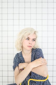 一位穿着蓝色圆点连衣裙的体贴金发少年，坐在瓷砖背景前