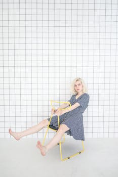 一位金发女子身穿蓝色连衣裙，赤脚坐在黄色椅子上，背景是白色瓷砖