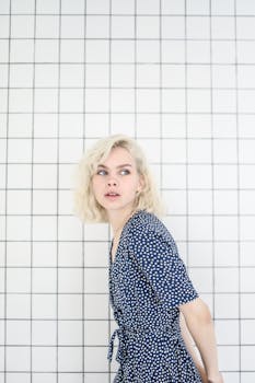 一位年轻的金发女子穿着蓝色圆点连衣裙，若有所思地站在瓷砖墙边