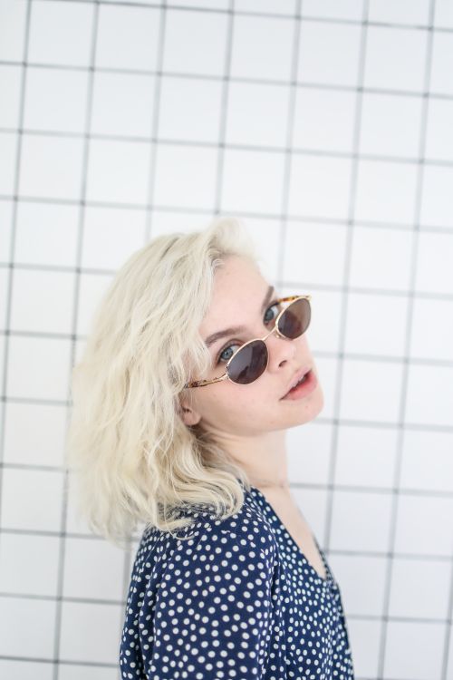 Free Portrait of a young woman with blonde hair and sunglasses against a tiled wall. Stock Photo