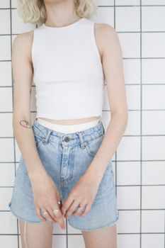 Teen girl in casual style wearing denim shorts and white top, standing against tiled background.