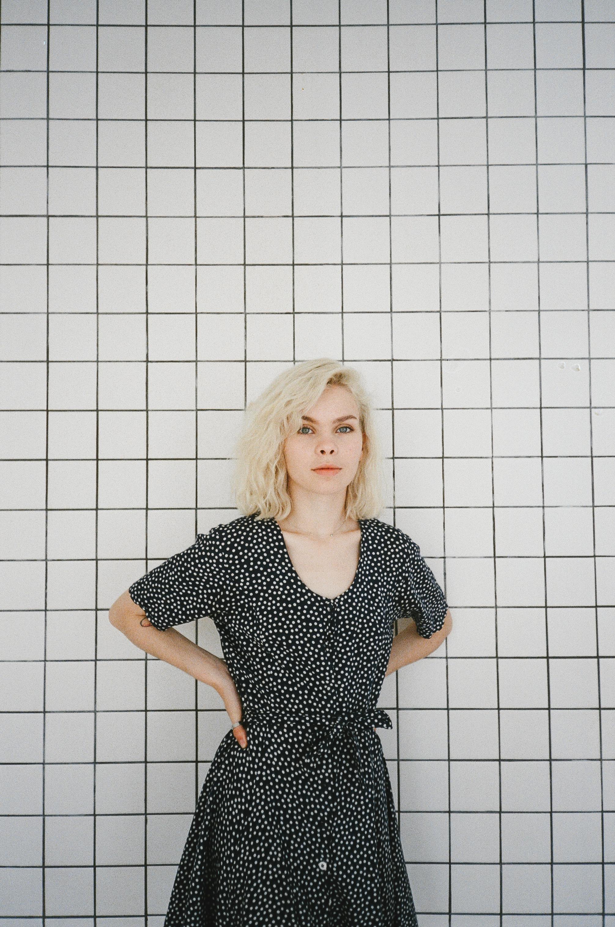 金发年轻女子穿着圆点连衣裙，自信地靠在网格图案的墙上摆出姿势