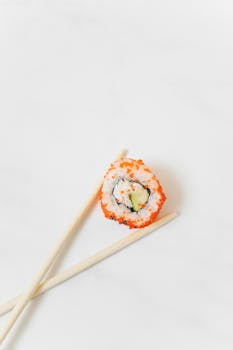 A close-up of a sushi roll with avocado and salmon, accompanied by chopsticks on a white background.