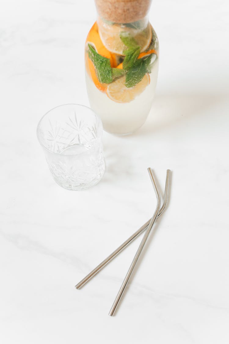 Fresh Water With Oranges And Mint In A Bottle, Crystal Drinking Glass And Metal Straws Lying On A Marble Counter 