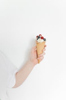 A hand holds an ice cream cone topped with fresh raspberries and blueberries on a white background.