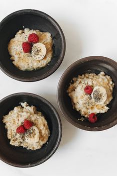 Top view of oatmeal bowls with banana, raspberries, and chia seeds on a white surface.