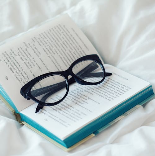 Free Eyeglasses placed on opened book on white bedsheets of soft bed in light bedroom Stock Photo