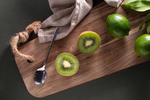 Sliced Kiwi Fruit Beside Limes and Silver Spoon on Brown Wooden Board