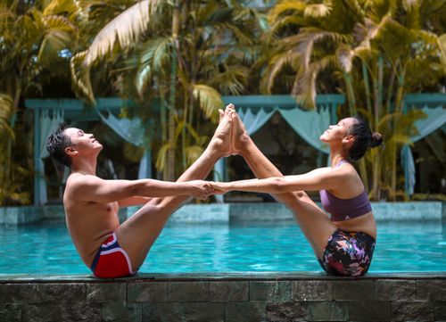 Free A couple performing yoga poses by a swimming pool surrounded by lush greenery. Stock Photo