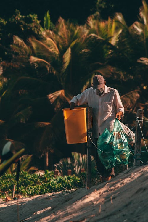 Unrecognizable black male in casual wear and cap collecting trash from street garbage bins in tropical country