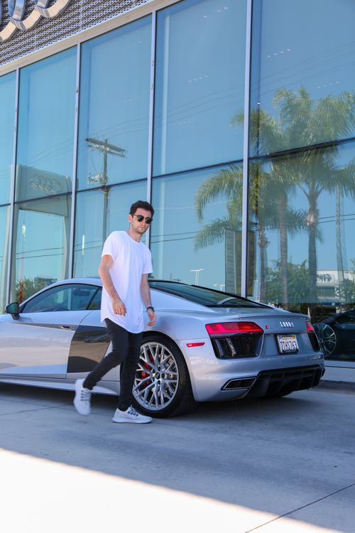 Free Full body of slender young male in casual clothing and sunglasses walking through street along luxury vehicle parked on pavement by building with broad transparent windows in daylight Stock Photo
