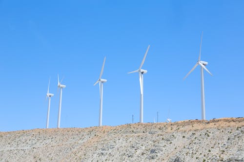 Vibrant blue cloudless sky over tall white wind turbines on slope in empty field in daytime