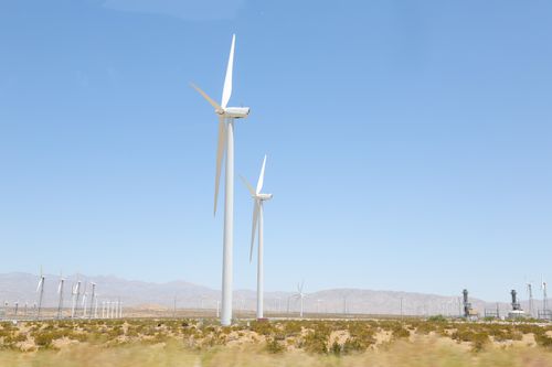 Free Tall wind energy converters with long steel towers and long thin rotor blades in meadow covered with tiny green bushes under light blue cloudless sky Stock Photo