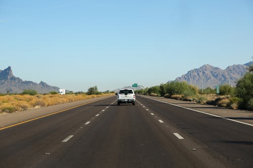 Light blue cloudless endless sky over white automobile riding on wide even paved road surrounded by bushes and mountain ridges
