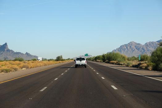 Light blue cloudless endless sky over white automobile riding on wide even paved road surrounded by bushes and mountain ridges