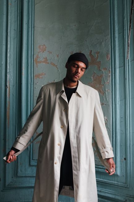 Fashion portrait of a man in a beige coat standing indoors against a rustic, textured wall.
