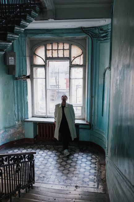 Man stands in vintage stairwell with elegant window, wearing beige coat. Natural light highlights shabby interior.