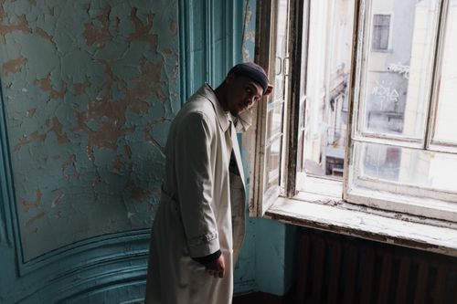 Free A man in a beige coat stands pensively by a vintage window in an old building with peeling paint. Stock Photo