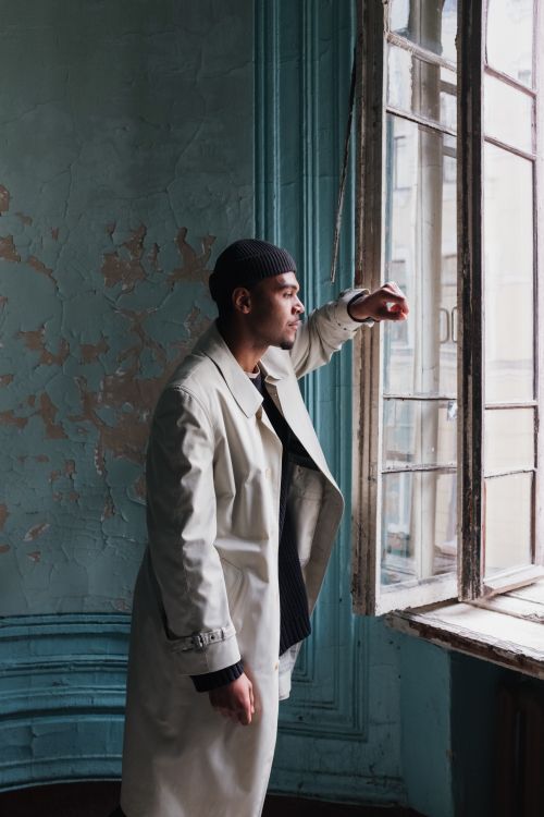 Free Man in a beige coat looks out a battered window in a shabby, vintage room. Stock Photo