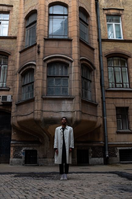A man stands in front of an aged, vintage building facade on a daylight street.
