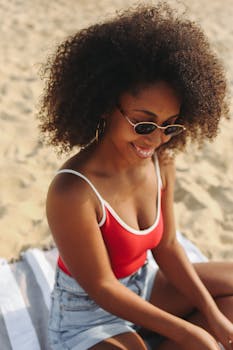 A cheerful woman in sunglasses enjoys a sunny day at the beach sitting on sand.