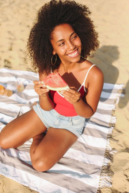Free Smiling woman with curly hair sitting on a beach, enjoying a juicy slice of watermelon under the sun. Stock Photo