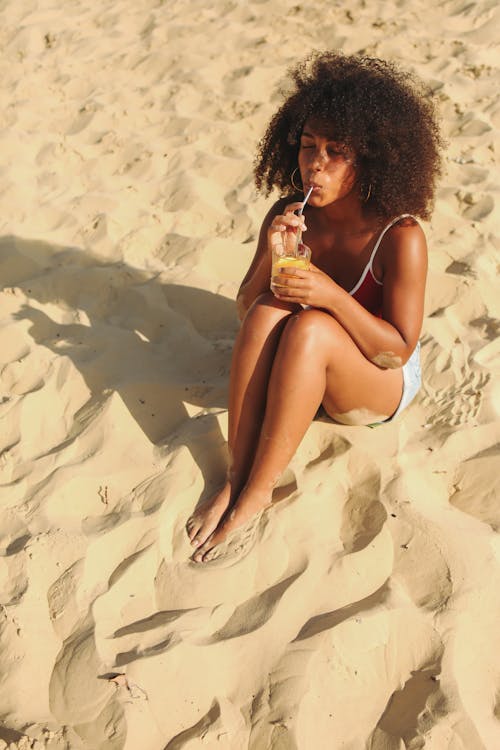 Free Woman Drinking on a Clear Glass While Sitting on White Sand Stock Photo