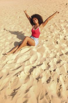 A happy woman with curly hair enjoying a sunny day at the beach, sitting with arms raised in delight.