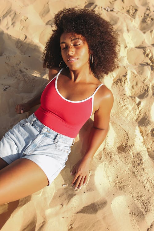 Free Woman in Red and White Spaghetti Strap Top and Denim Shorts Lying on Beach Sand Stock Photo