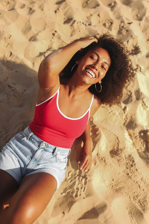 Free Smiling Woman in Red and White Spaghetti Strap Top and Denim Shorts Lying on Beach Sand Stock Photo