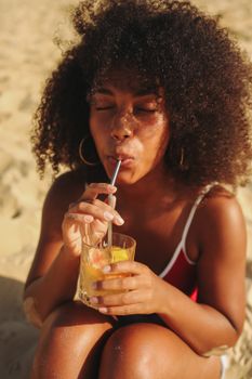 Sun-kissed woman with afro hair sipping a refreshing drink on the beach, eyes closed in relaxation.