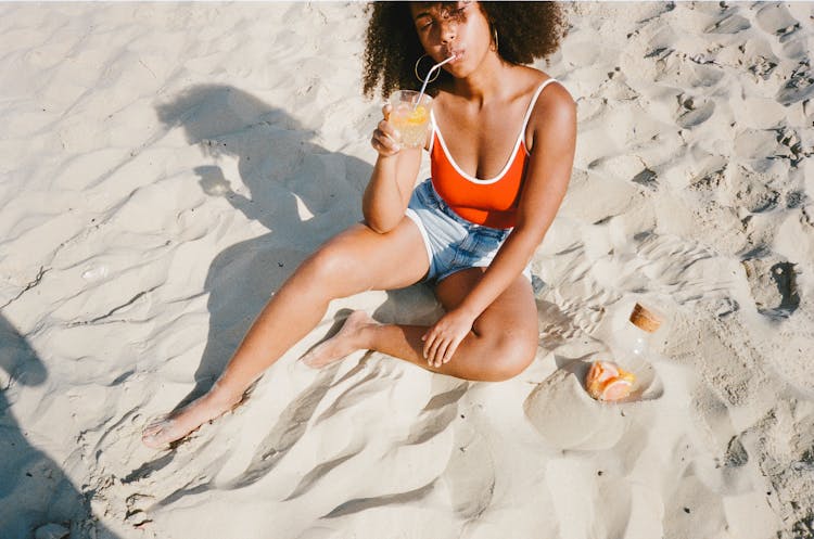 Woman Sitting On Sand Drinking A Glass Of Refreshment