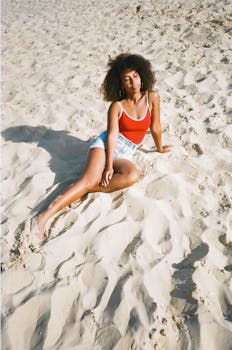 Woman sitting on a sandy beach in a red top and denim shorts, enjoying the sun.