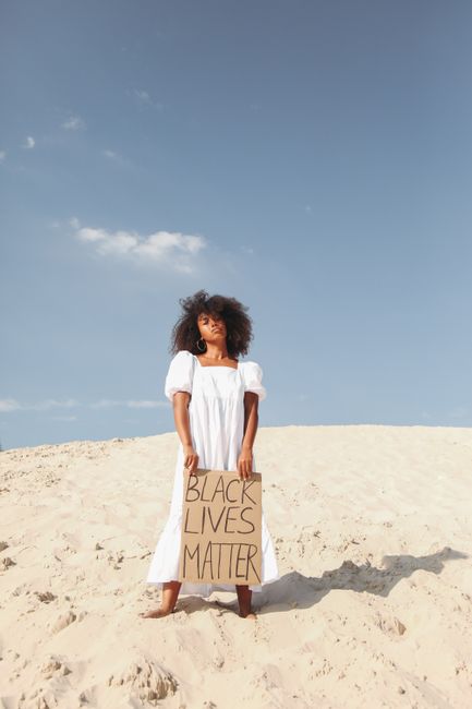 Black woman standing in desert holding a 'Black Lives Matter' sign under clear sky.