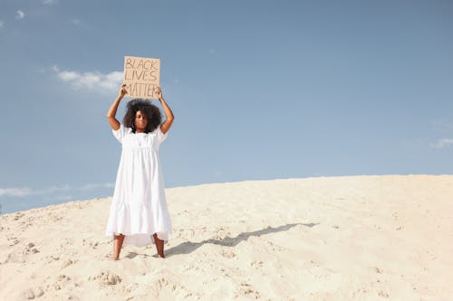 Woman Holding Poster in White Dress Standing on Sand