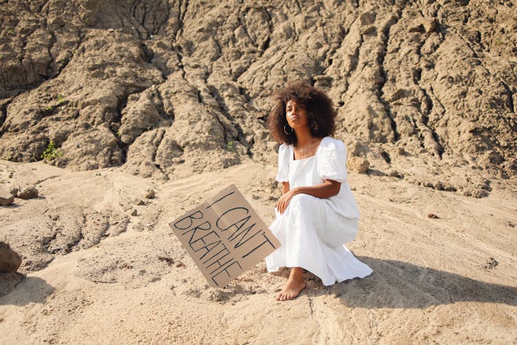 A Woman In White Dress Sitting On The Ground Beside A Cardboard With Message