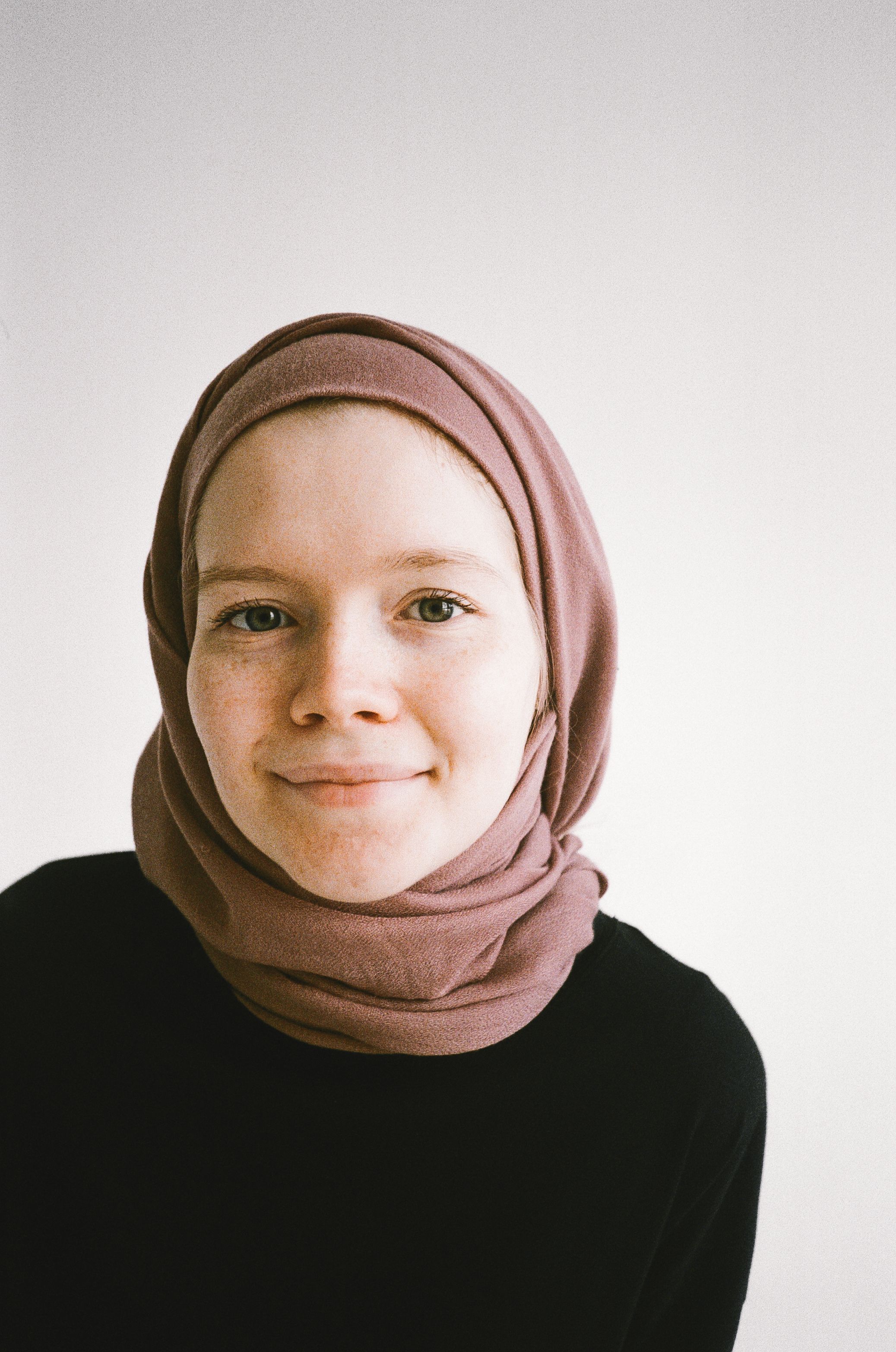 A close-up portrait of a smiling woman wearing a pink hijab with a neutral background.
