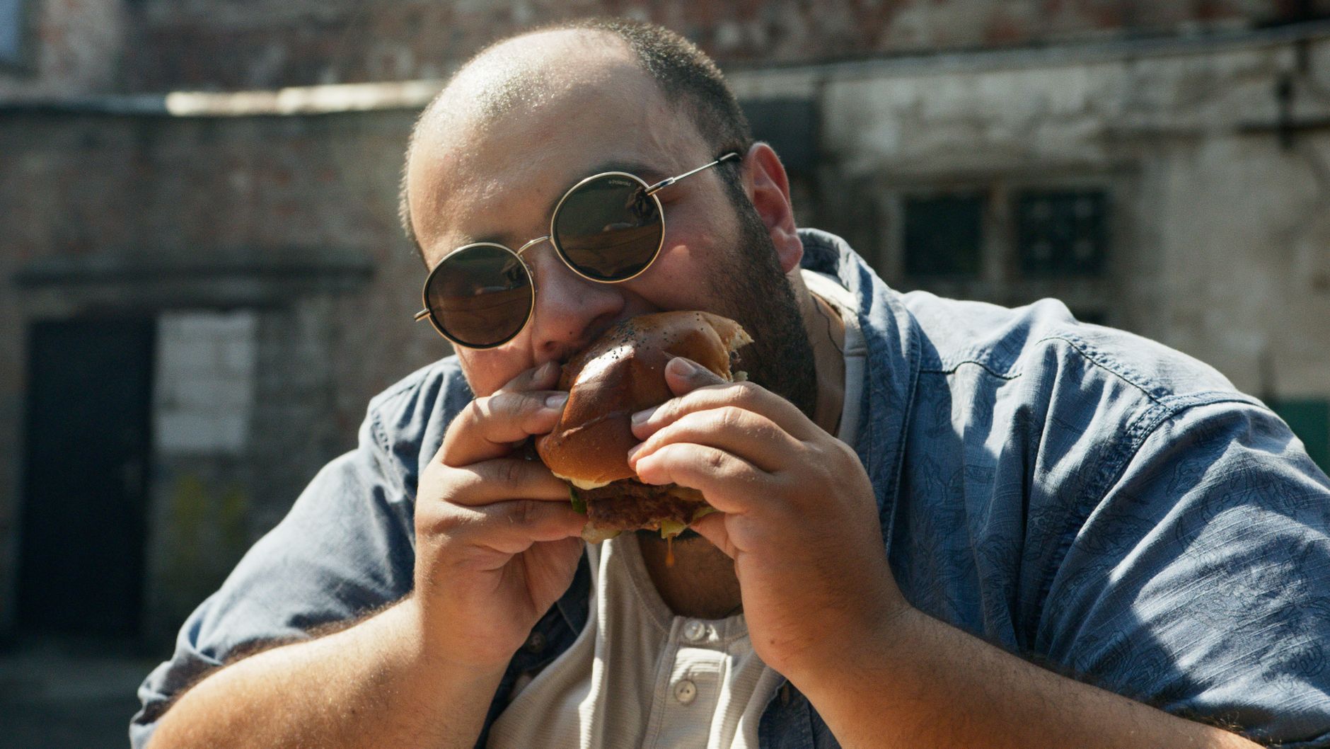 A man wearing sunglasses enjoys a big burger outdoors in London.
