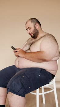 A plus size man with facial hair sits on a chair indoors, focused on using his smartphone, against a beige background.