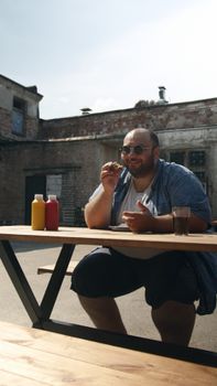 Smiling man in sunglasses enjoys food outdoors at a picnic table.