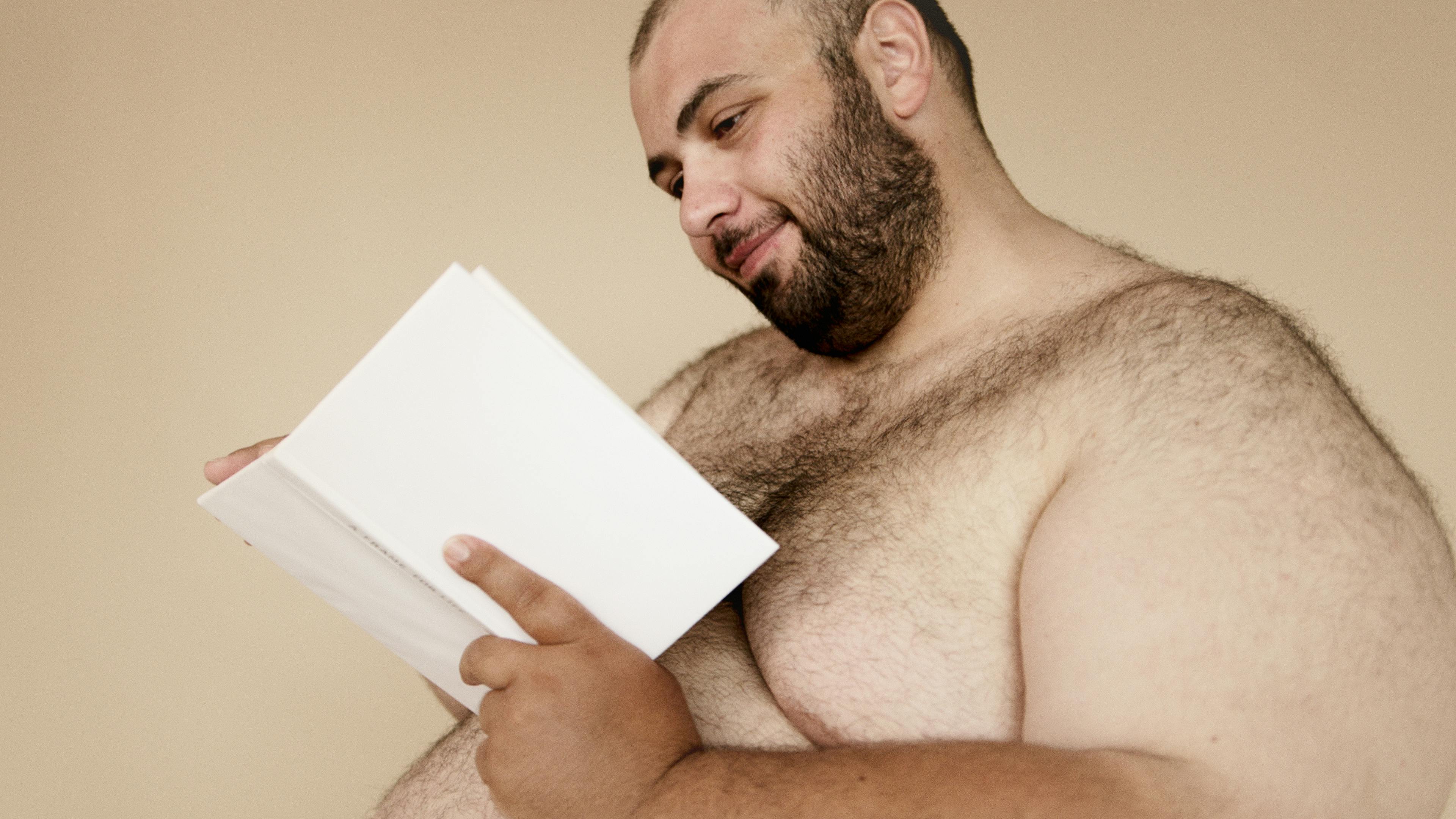 A bearded shirtless man smiling while reading a book against a neutral background.