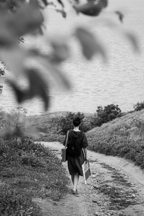 Black and white back view female wearing casual dress strolling along rural pathway on grassy hill slope towards calm sea