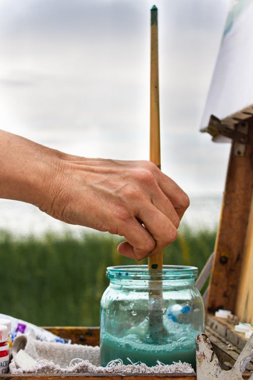 Crop faceless female artist cleaning paintbrush in dirty water in glass jar while drawing in grassy nature