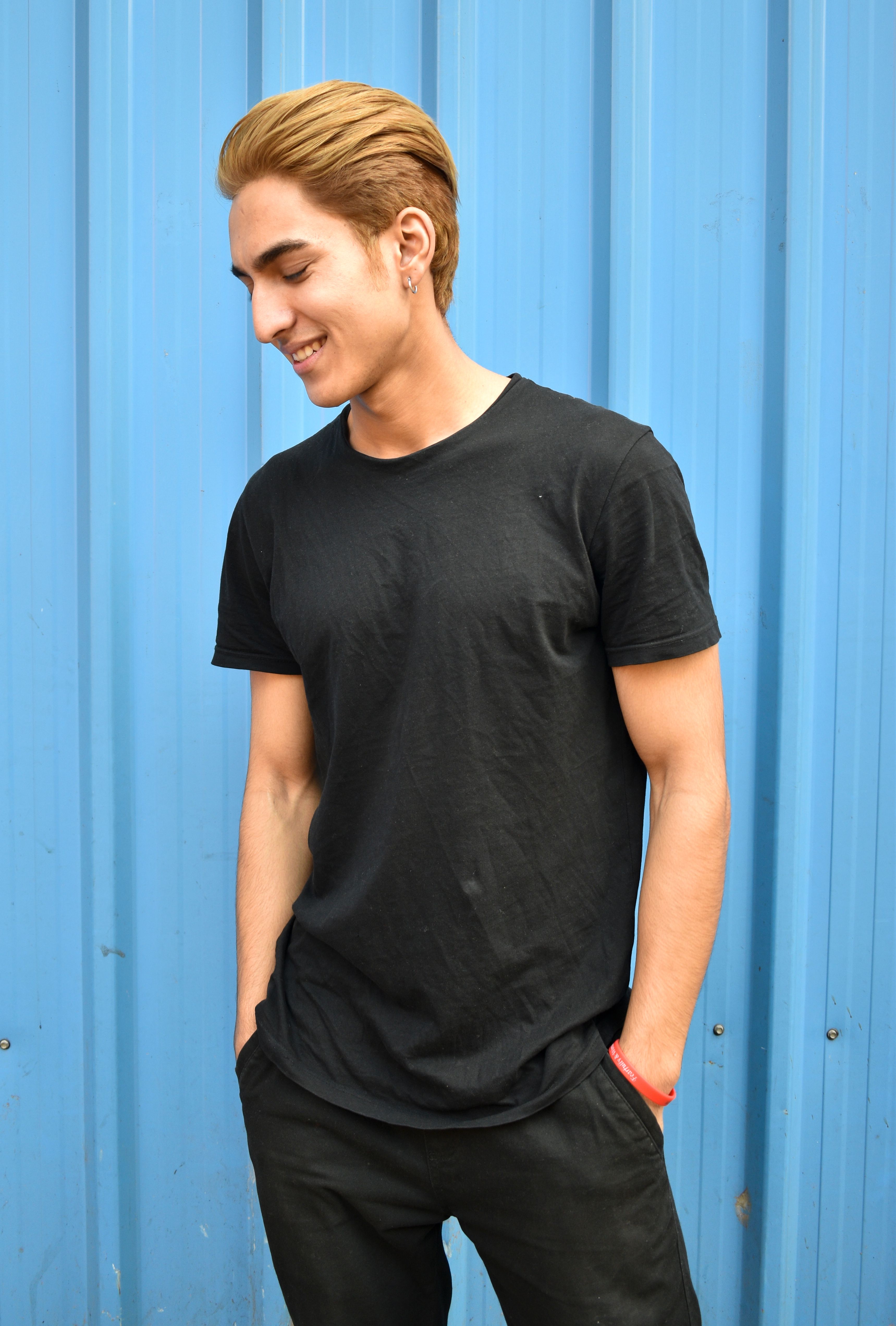 A young man stands smiling against a vibrant blue wall, wearing a black t-shirt and exuding a relaxed vibe.